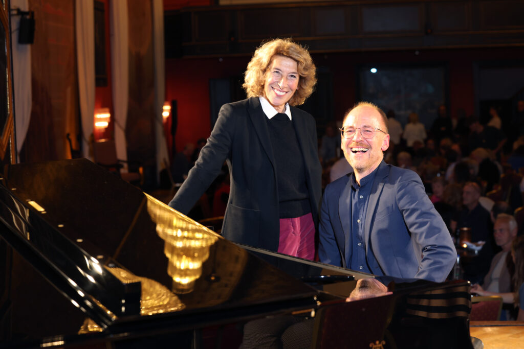 Adele Neuhauser Foto Sigi Muller www.augenblick-fotografie.com (9) - IN München - Das Stadtmagazin Two performers at a grand piano smiling for the audience in a dimly lit venue.