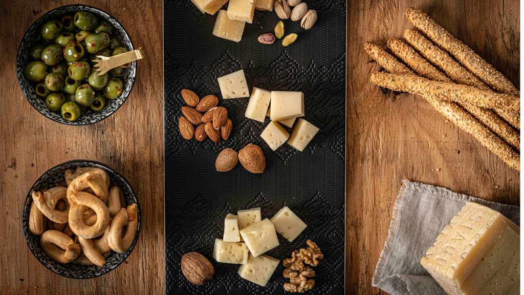 Asiago-2 - IN München - Das Stadtmagazin Assorted cheeses, olives, nuts, and sesame breadsticks arranged on a rustic wooden table with two bowls of snacks and a black central board.