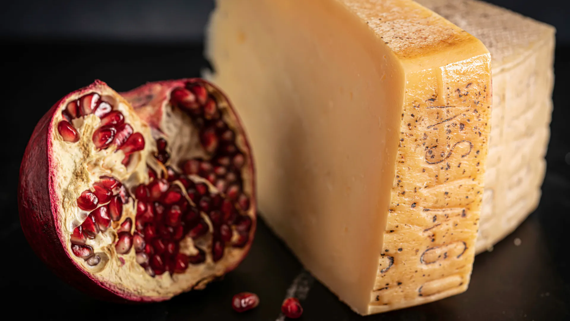 Pomegranate half with bright red seeds next to a large block of yellow cheese on a dark surface.