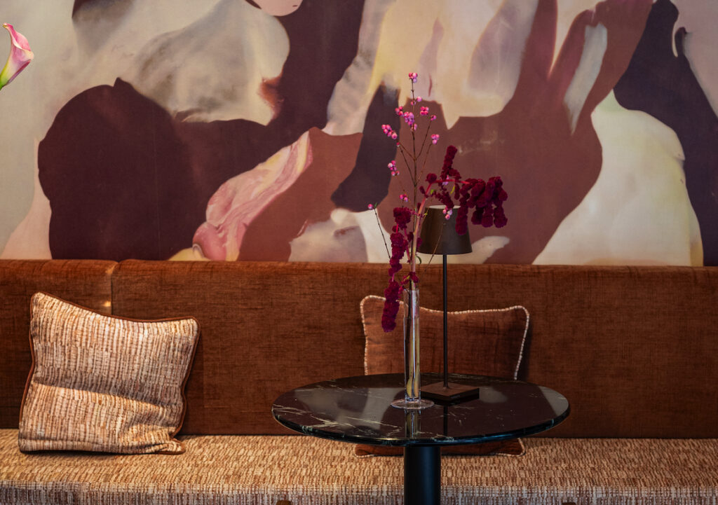 Screenshot - IN München - Das Stadtmagazin Brown sofa with beige patterned cushions, a round black marble table, and a vase of purple flowers against an abstract mural.