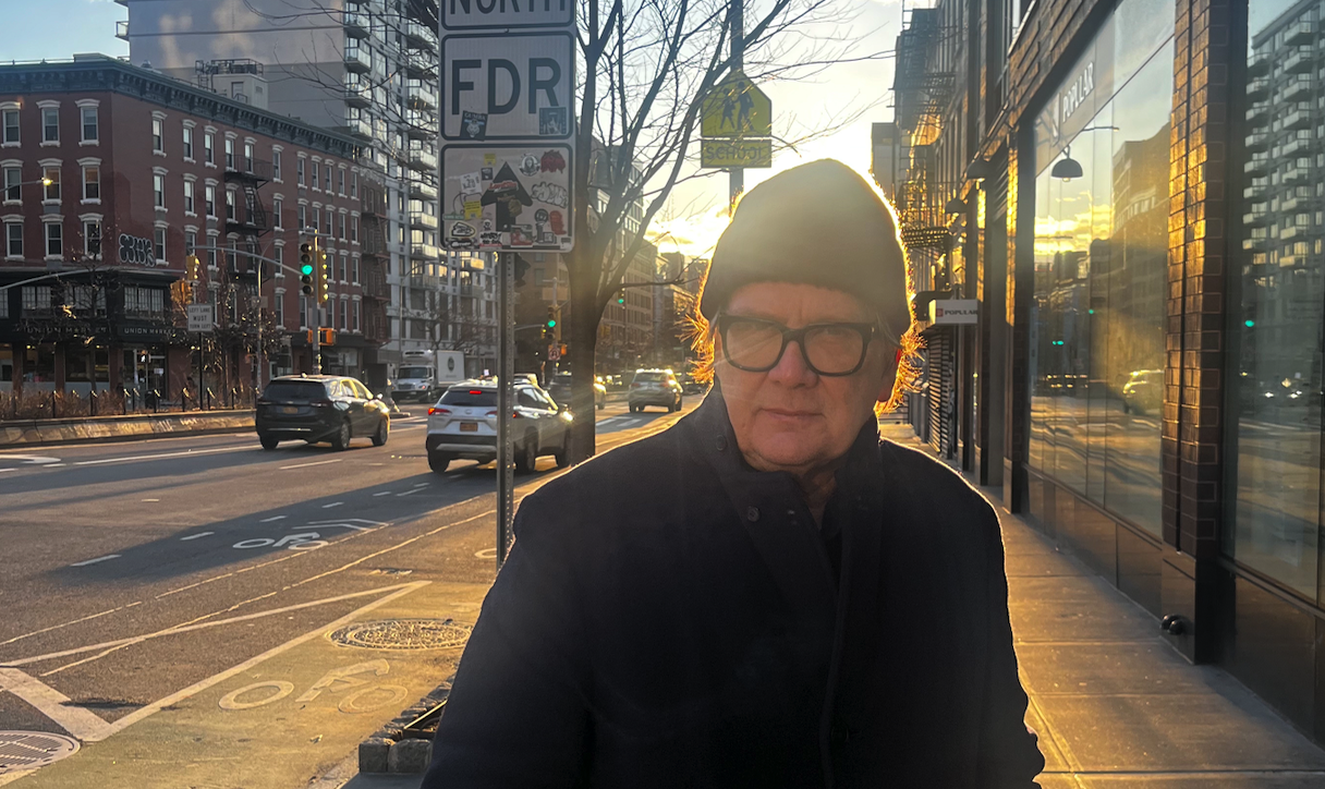 Older man in a black coat and beanie walking along a city sidewalk at sunset, storefronts and traffic in the background.
