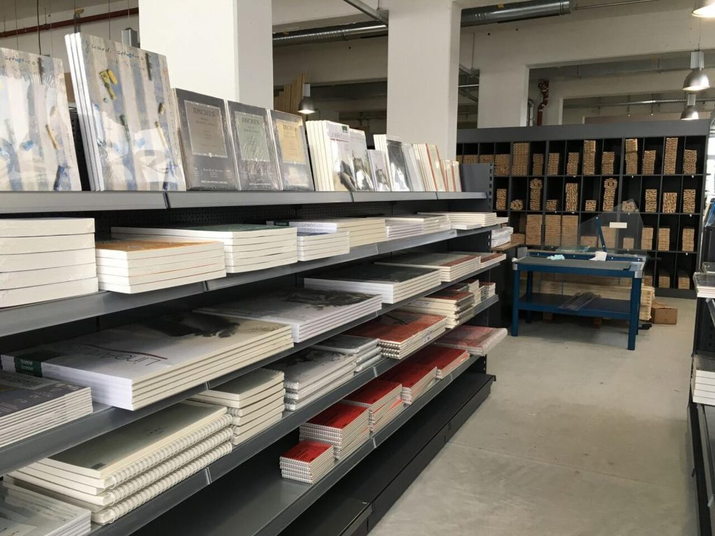 Boesner - IN München - Das Stadtmagazin Store aisle with metal shelves holding stacked notebooks and art prints; a blue worktable is visible in the background with wooden storage cubes along the back wall.