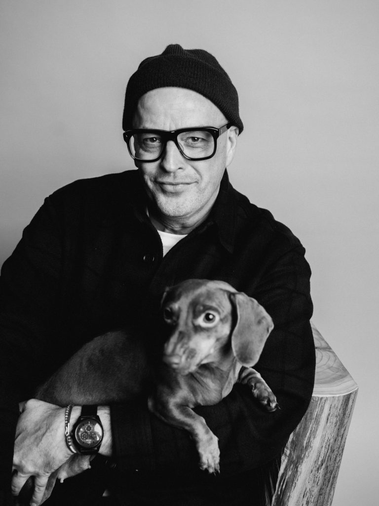 _DSF7218 - IN München - Das Stadtmagazin Man wearing a beanie and glasses holding a small dog on his lap, in a black-and-white portrait.