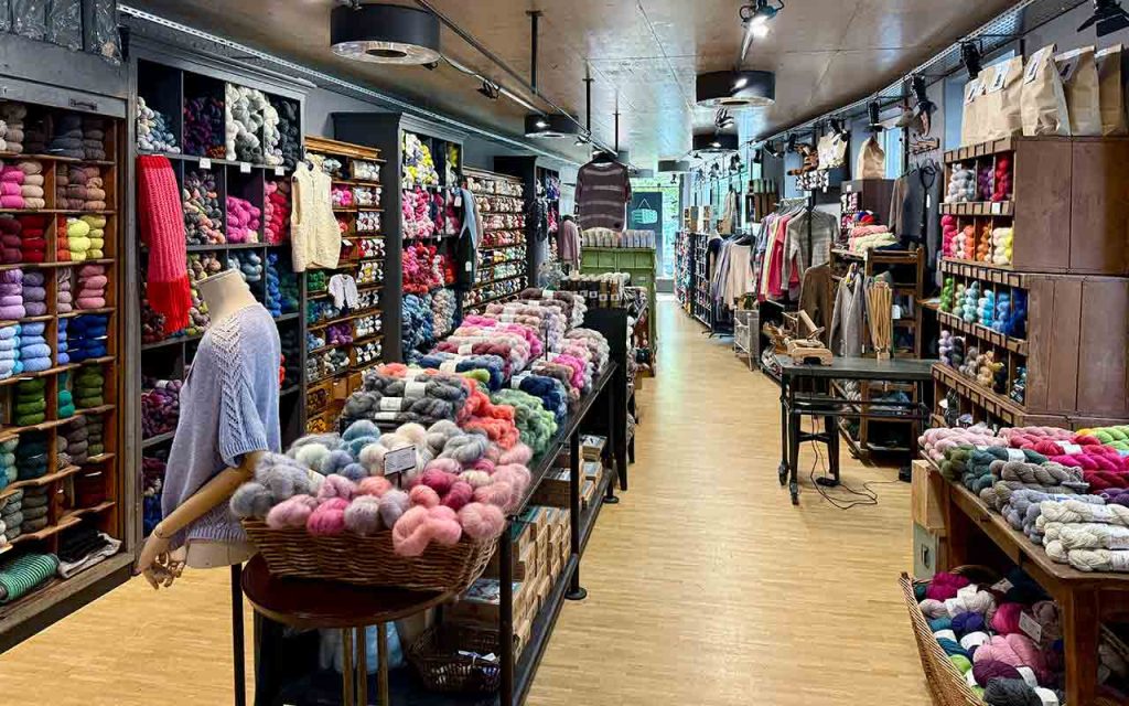 Die_Mercerie_Laden_2025_1920x1920 - IN München - Das Stadtmagazin Colorful yarn shop interior with tall shelves of skeins, baskets of fluffy yarn, knitting mannequins, and a long central aisle.