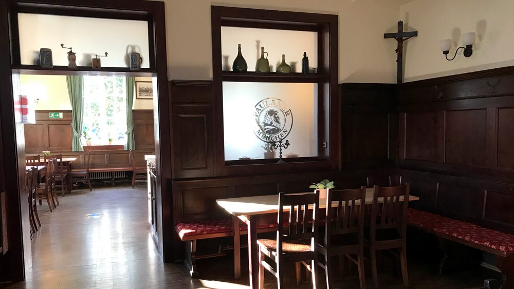 Wood-paneled dining room with a long central table, chairs, and bench seating in a sunlit, cozy restaurant.