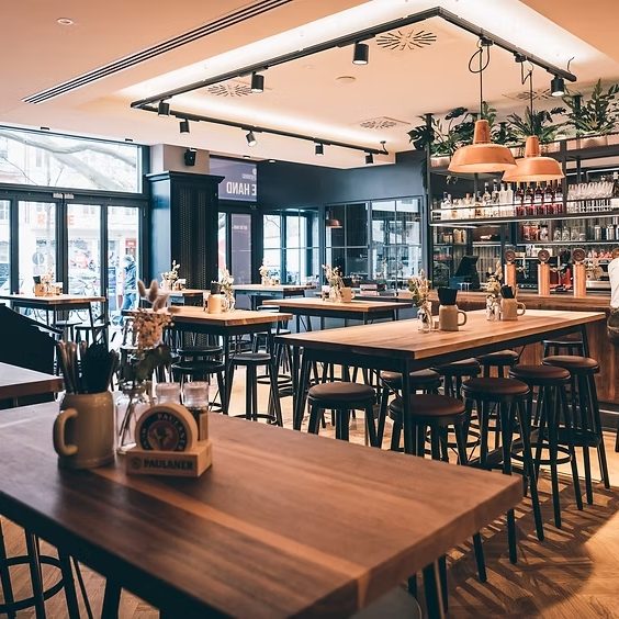 Bright, modern cafe interior with wooden tables and stools, hanging copper lamps, plants, and a bar along the back wall.
