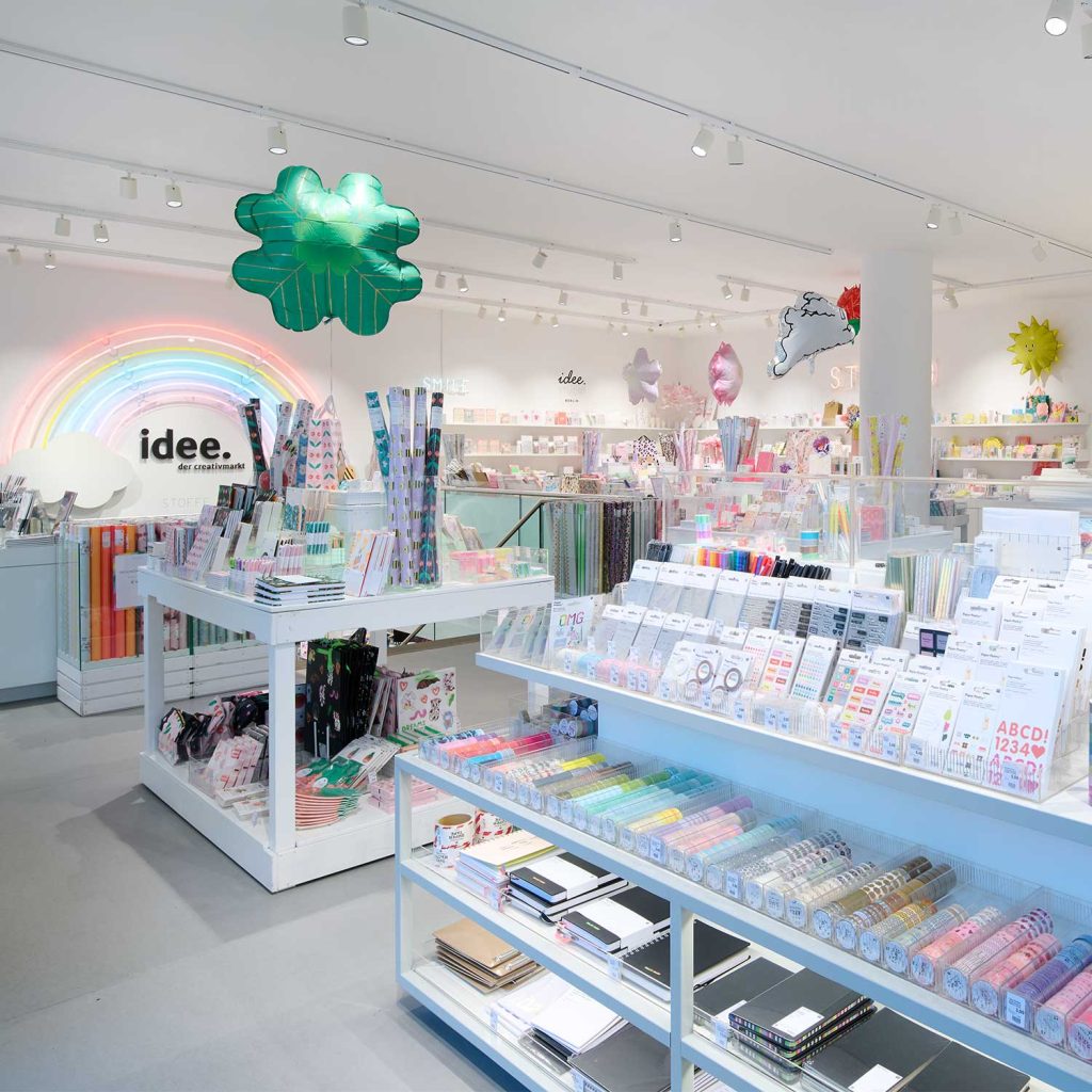 Bright stationery store with white shelves filled with colorful notebooks and rolls of decorative paper; neon rainbow display in back and oversized green shamrock balloon overhead.