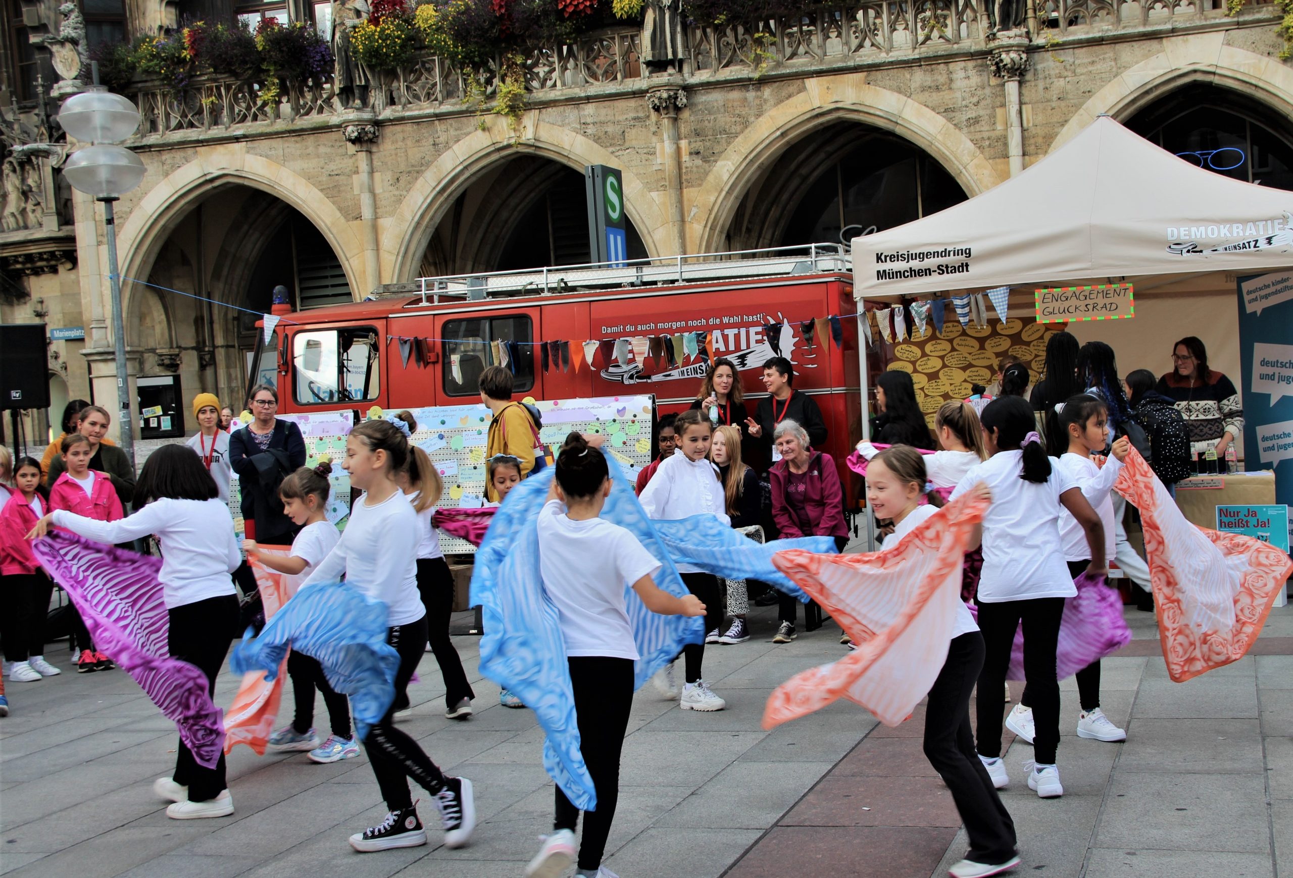 Group of children and adults dancing with blue, pink, and orange scarves in a city square, with a red vehicle and a tent behind them.