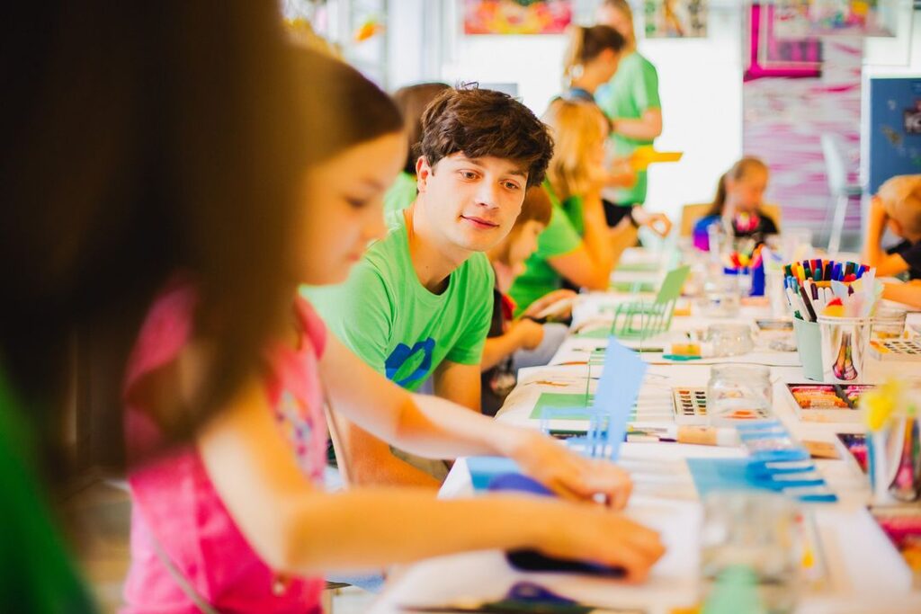 Little Art - IN München - Das Stadtmagazin Group of children at a long craft table, painting and coloring with colorful supplies in a bright classroom.