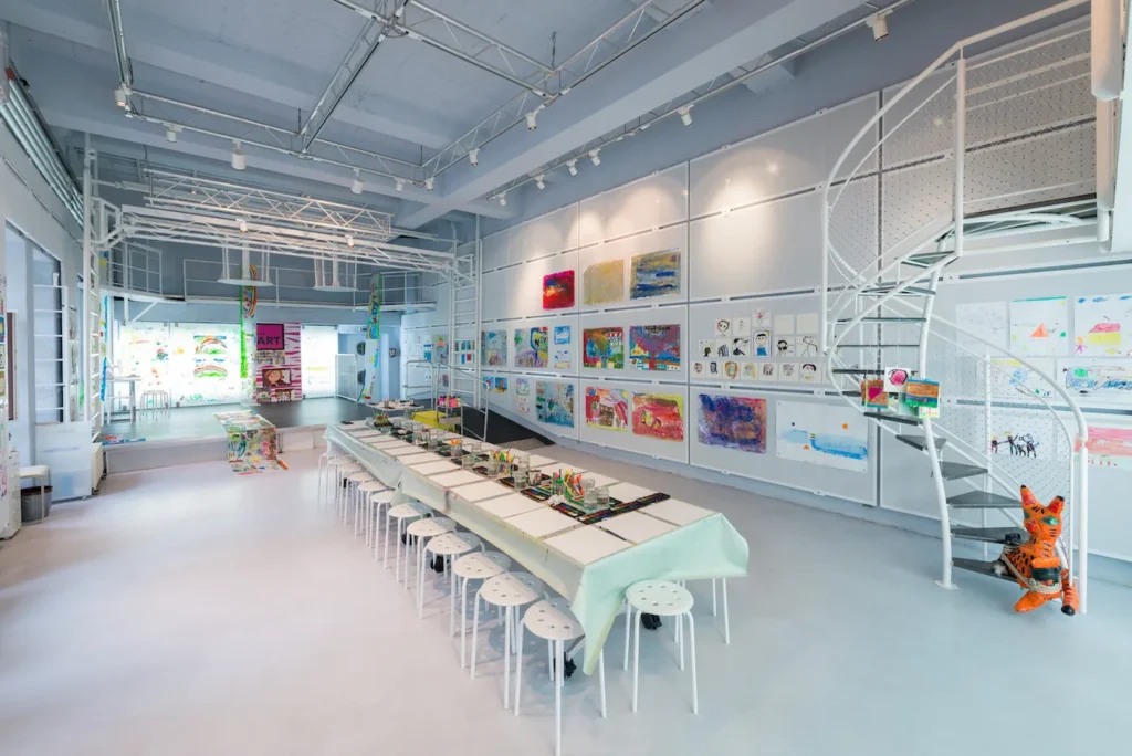 Little Art Org München - IN München - Das Stadtmagazin Bright art studio with a long craft table, white stools, and walls filled with colorful drawings and paintings.
