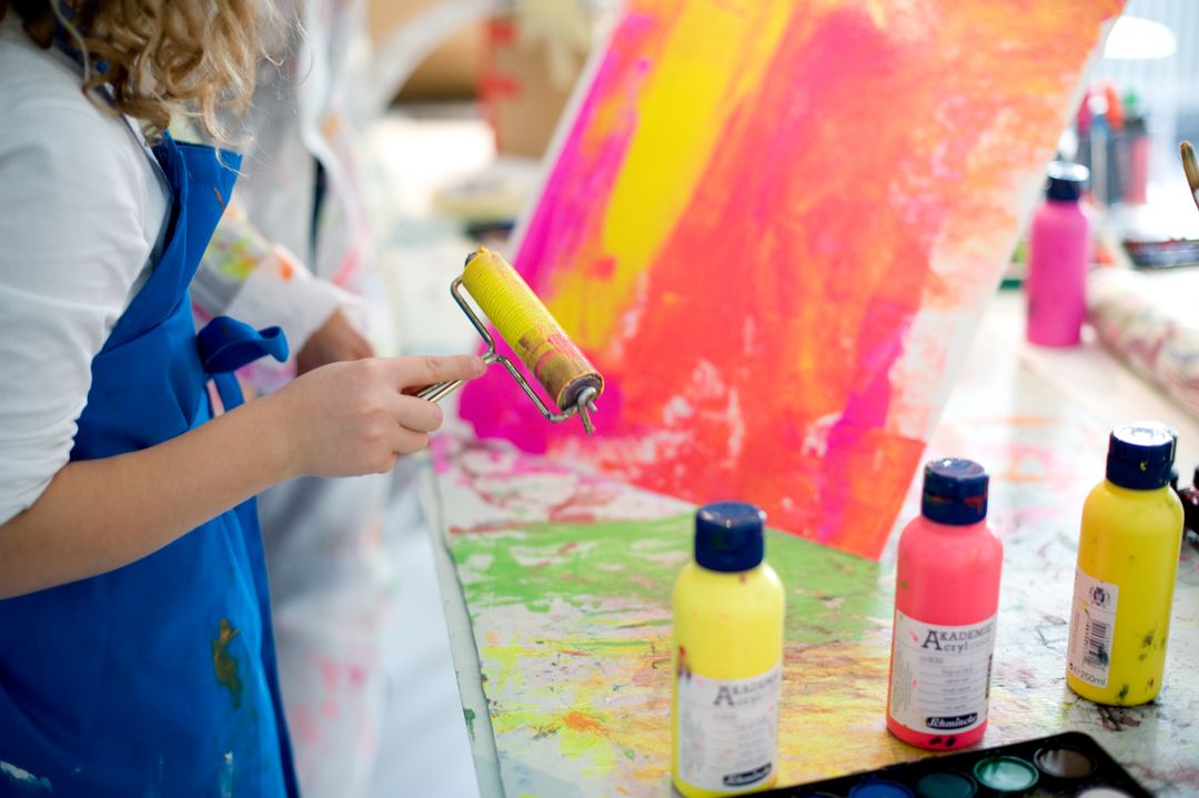 Person in a blue apron rolling yellow paint onto a vibrant abstract canvas with bright pinks and oranges nearby on a paint-splattered table.