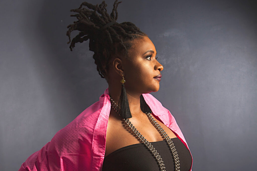 Profile of a woman with dreadlocks wearing a pink jacket, black top, and long beaded necklace against a dark blue background.
