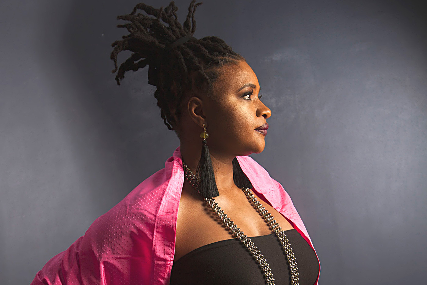 Profile of a woman with dreadlocks wearing a pink jacket, black top, and long beaded necklace against a dark blue background.