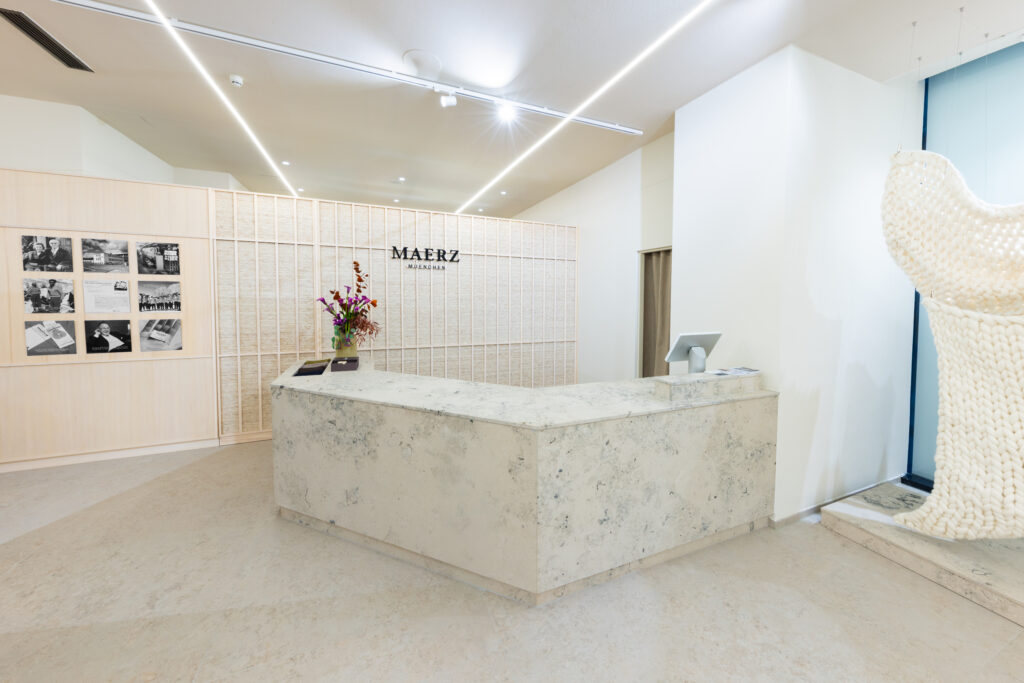 MAERZ_Muenchen_store_MUC_02 - IN München - Das Stadtmagazin Bright reception desk in a minimalist lobby, with a marble counter, a pale wood paneled wall of photos, and a bouquet of flowers in front.