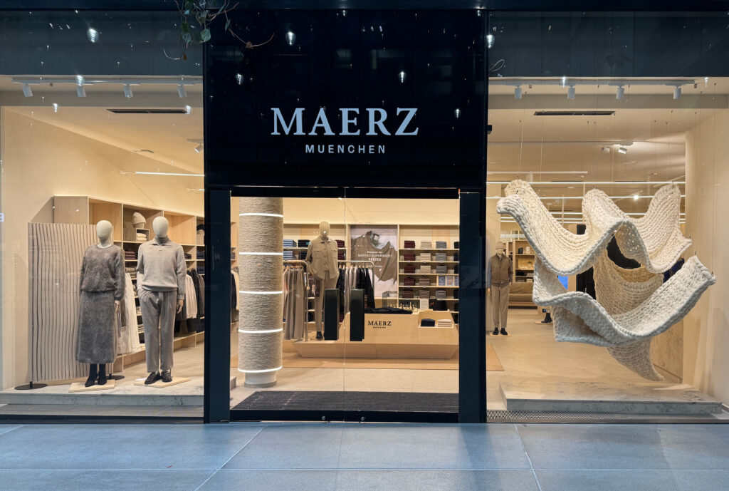 MAERZ_Muenchen_store_MUC_window_01 - IN München - Das Stadtmagazin Glass storefront of MAERZ MÜNCHEN clothing store with mannequins in neutral-toned outfits visible inside and a large knit sculpture to the right