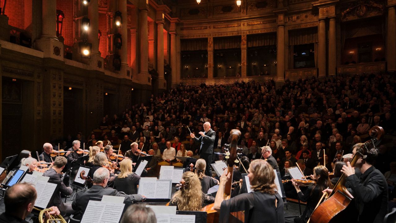 Orchestra performing on a grand concert hall stage, conductor leading musicians with an audience watching from the seats.