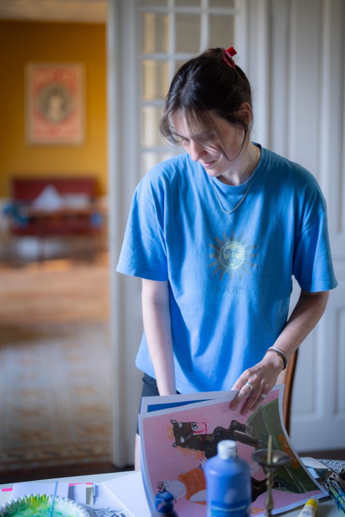 Minimalere_Phoebefongphotography - IN München - Das Stadtmagazin Young woman in a blue t-shirt looking down at colorful posters on a table indoors, arranging artwork.