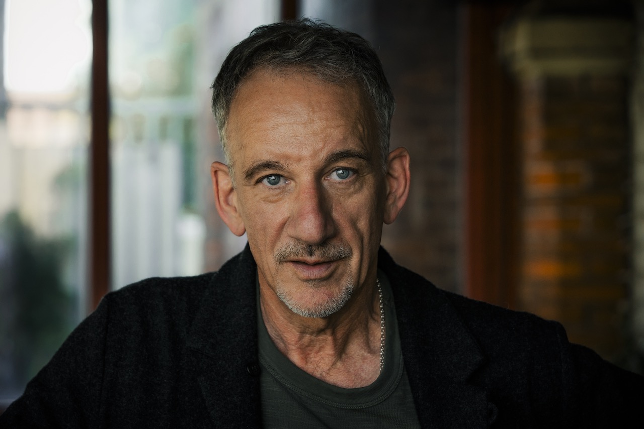 Portrait of a mature man with blue eyes and short gray hair, wearing a dark jacket indoors by a brick wall.