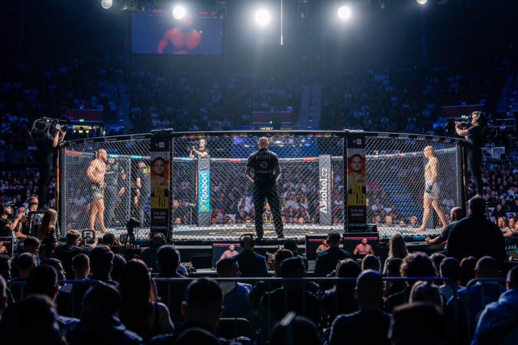 Wide shot of a mixed martial arts cage fight in a crowded arena, fighters inside the octagonal fence with a referee nearby and cameras filming from outside the cage.
