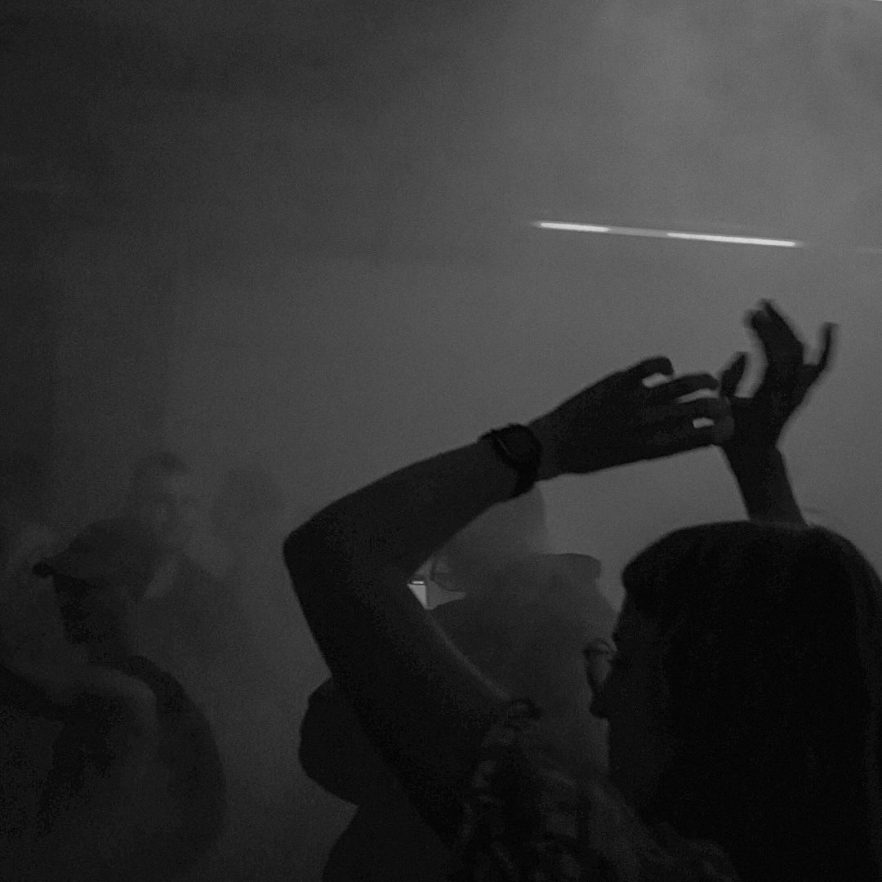 Silhouetted crowd dancing in a smoky, dark venue with hands raised in the air.
