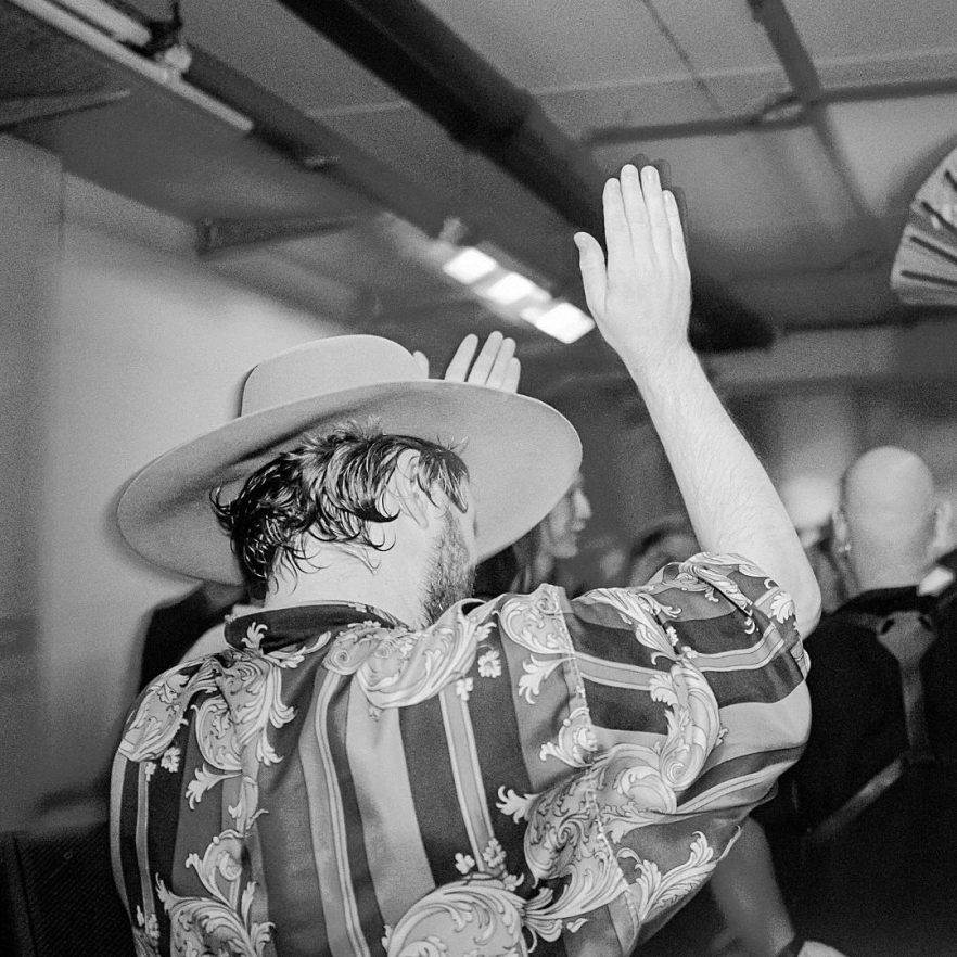 Man wearing a wide-brimmed hat and patterned shirt raises his hand at a crowded indoor party or club.
