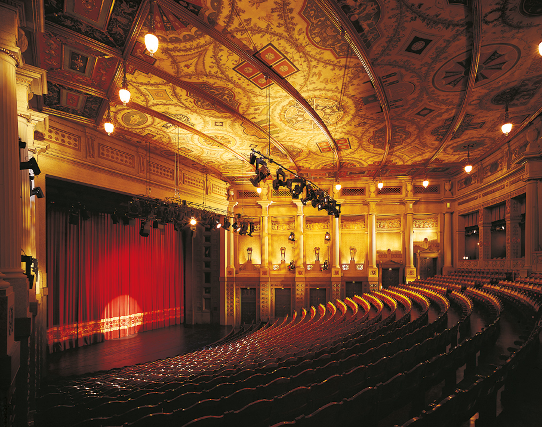 Ornate theater interior with a red stage curtain, gold-painted ceiling, and rows of empty seats.