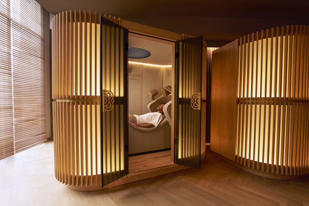 Curved wooden privacy pods in a spa, with a person reclining in a treatment chair inside one pod, warm lighting, and a large circular ceiling shower above.