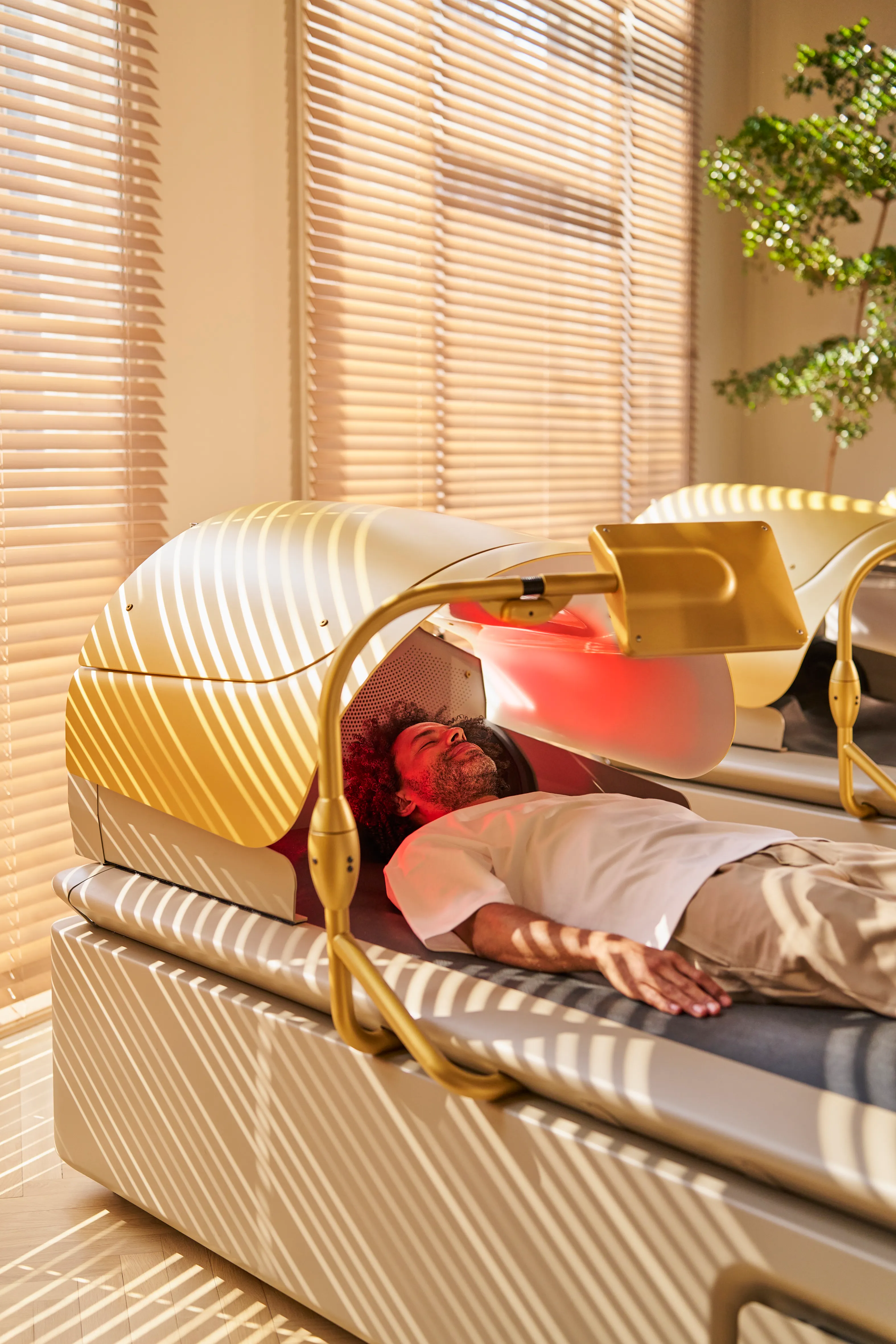Person resting inside a gold-trimmed curved spa bed in a sunlit room with wooden blinds behind.