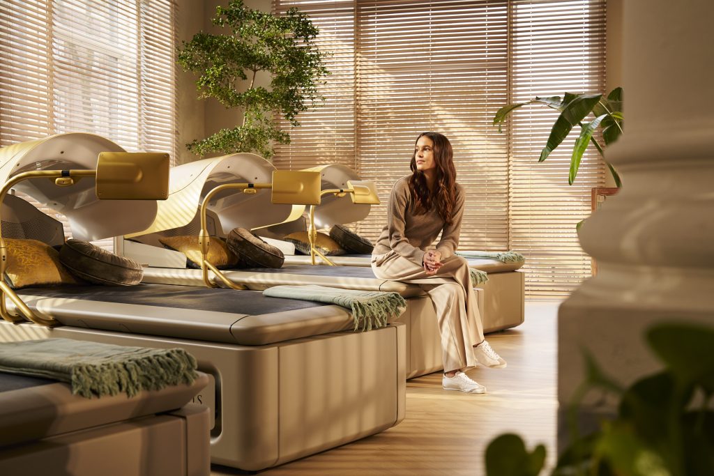 Woman sits on a beige lounge bed in a sunlit spa beside row of gold-framed tanning beds, with green plants and wooden blinds behind her, looking off to the side to the right.