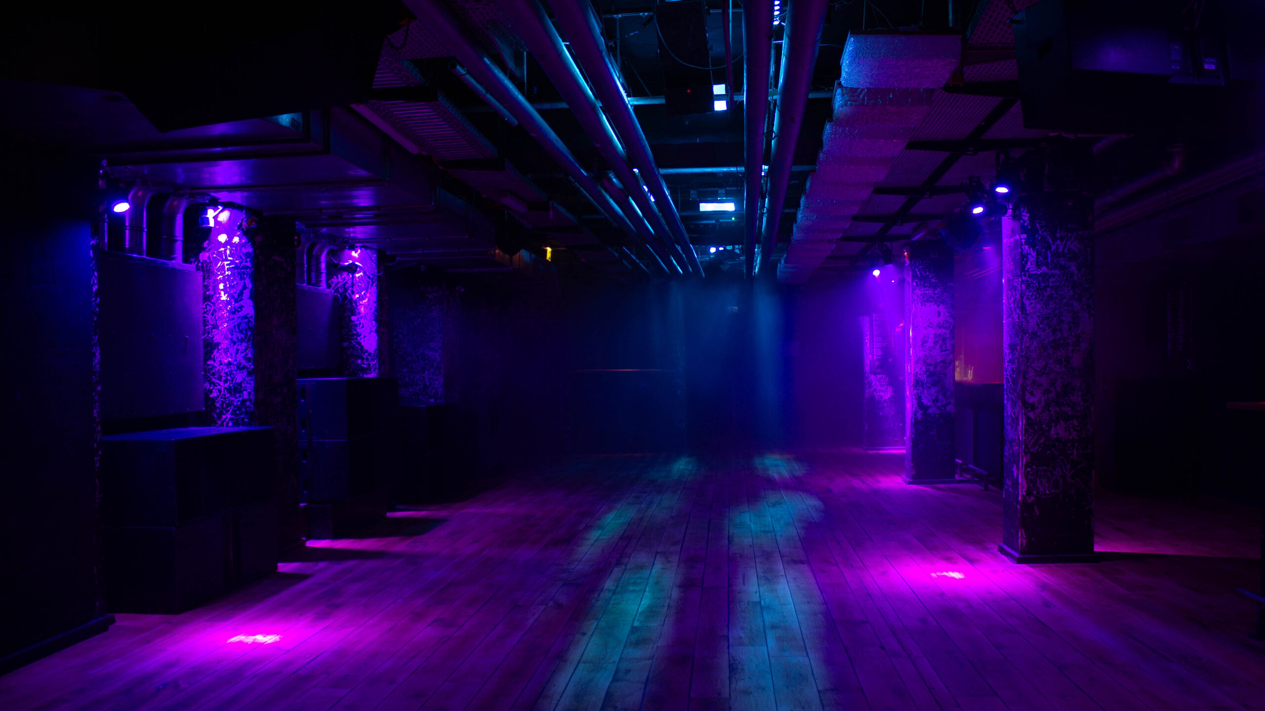 Rote-Sonne-Club-Foto-2-Lilith-Elea - IN MĂĽnchen - Das Stadtmagazin Wide shot of a dark nightclub with purple/blue lighting, concrete pillars, and a wooden floor, empty dance area left and right edge.