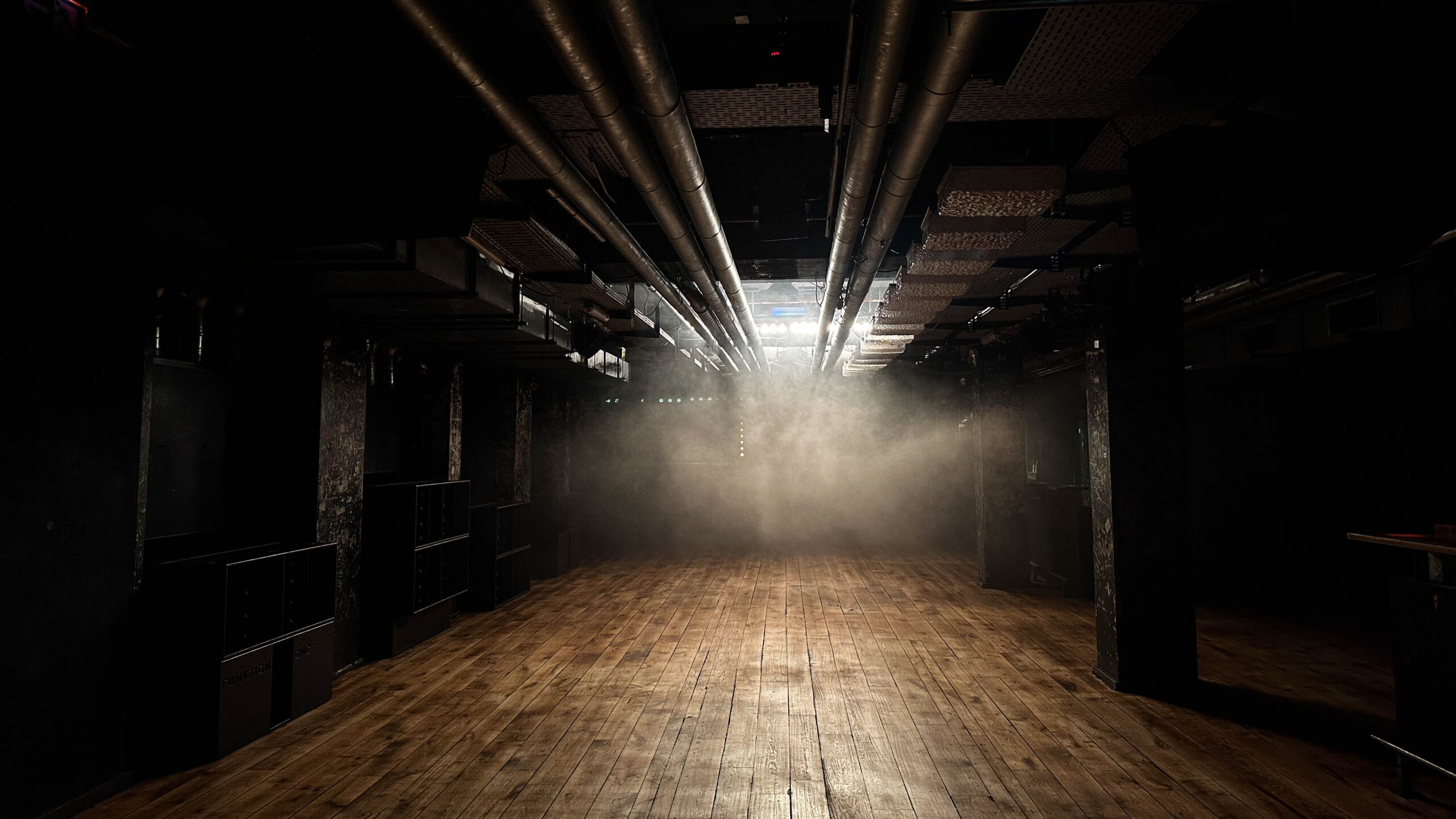 Rote-Sonne-Club-Foto-3-Lilith-Elea - IN MĂĽnchen - Das Stadtmagazin Dark, empty industrial-style theater with wooden stage and exposed pipes overhead, illuminated by bright backlights and hanging stage lights, with fog near the far end.