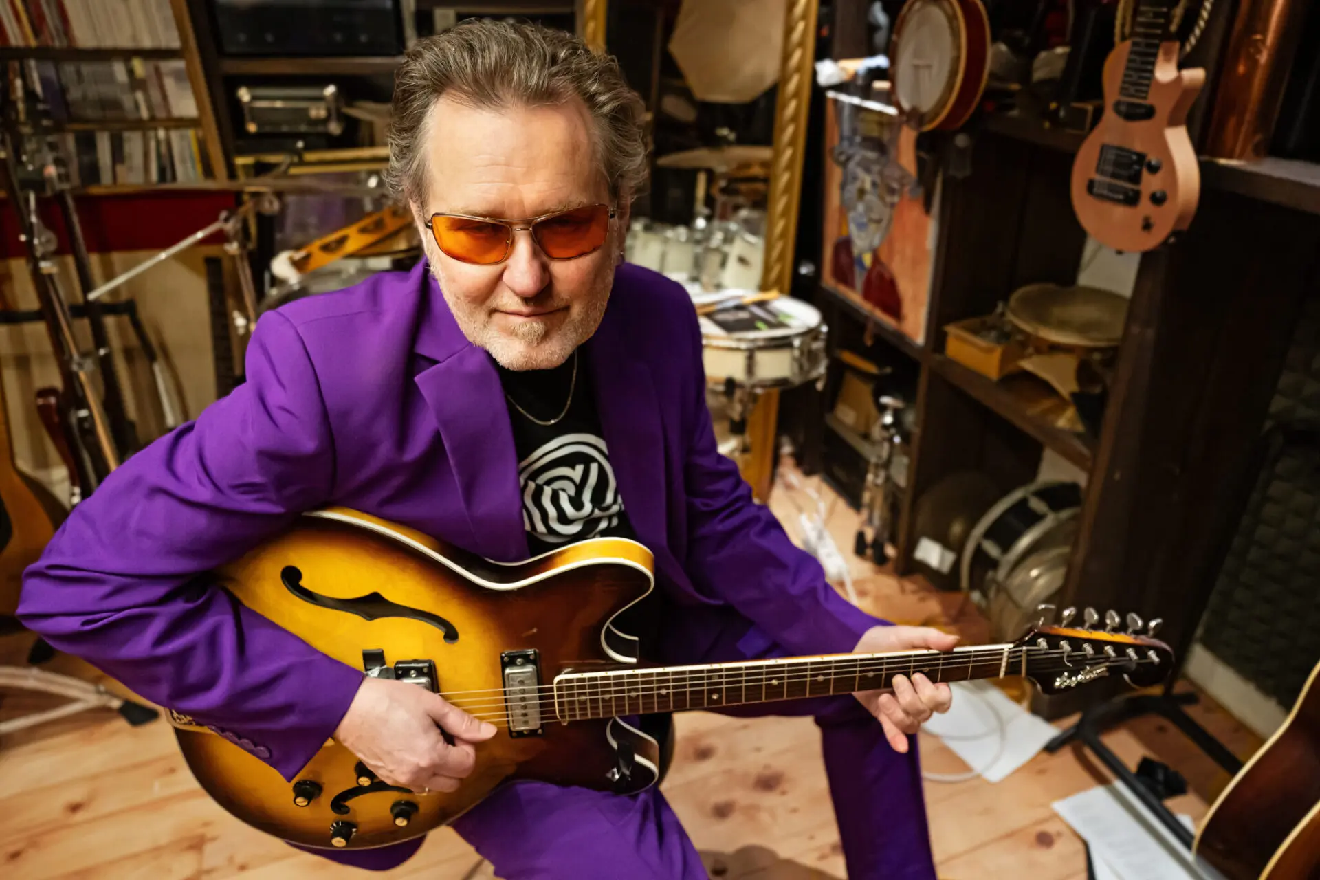 Musician wearing a purple suit and orange glasses playing a sunburst semi-hollow guitar in a cluttered music studio filled with guitars and gear