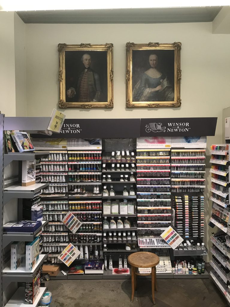 Two framed classical portraits in ornate gold frames mounted on a pale wall above art supplies in a shop setting.