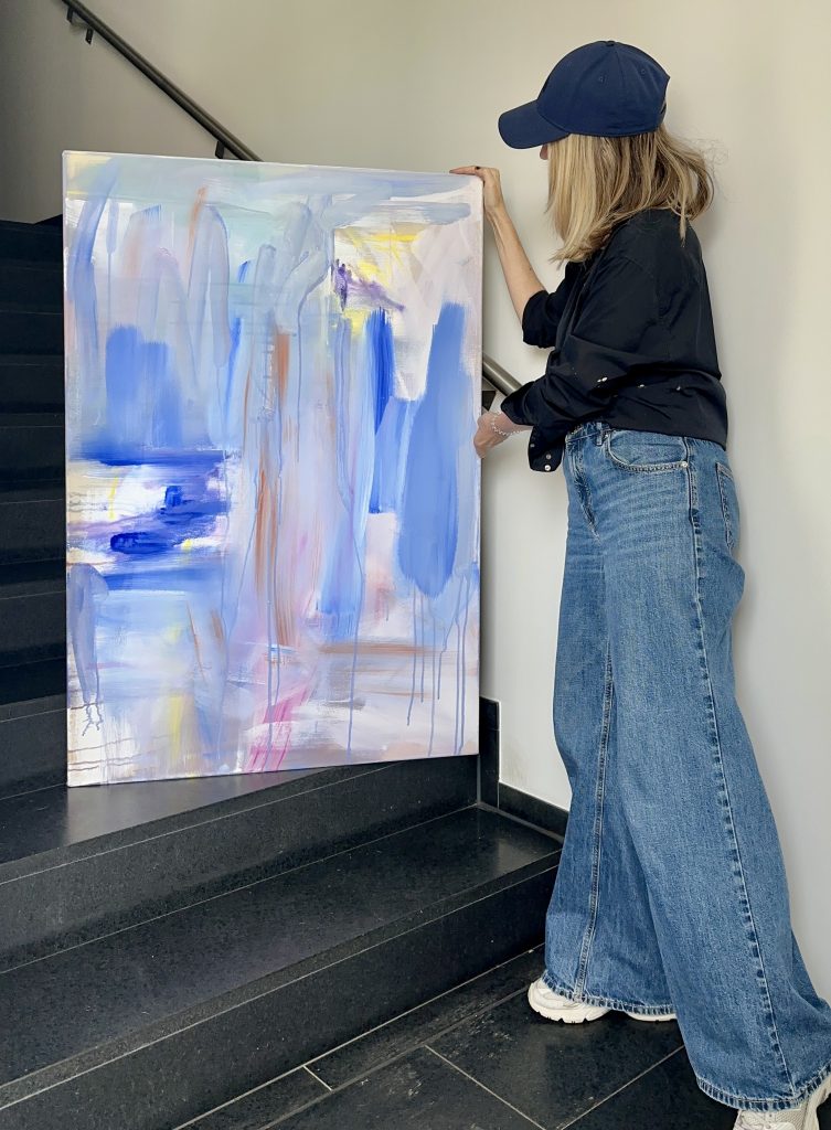 Person wearing a blue cap and jeans holds a large abstract painting on a staircase, with blue, lavender, and yellow brushstrokes in soft pastels.