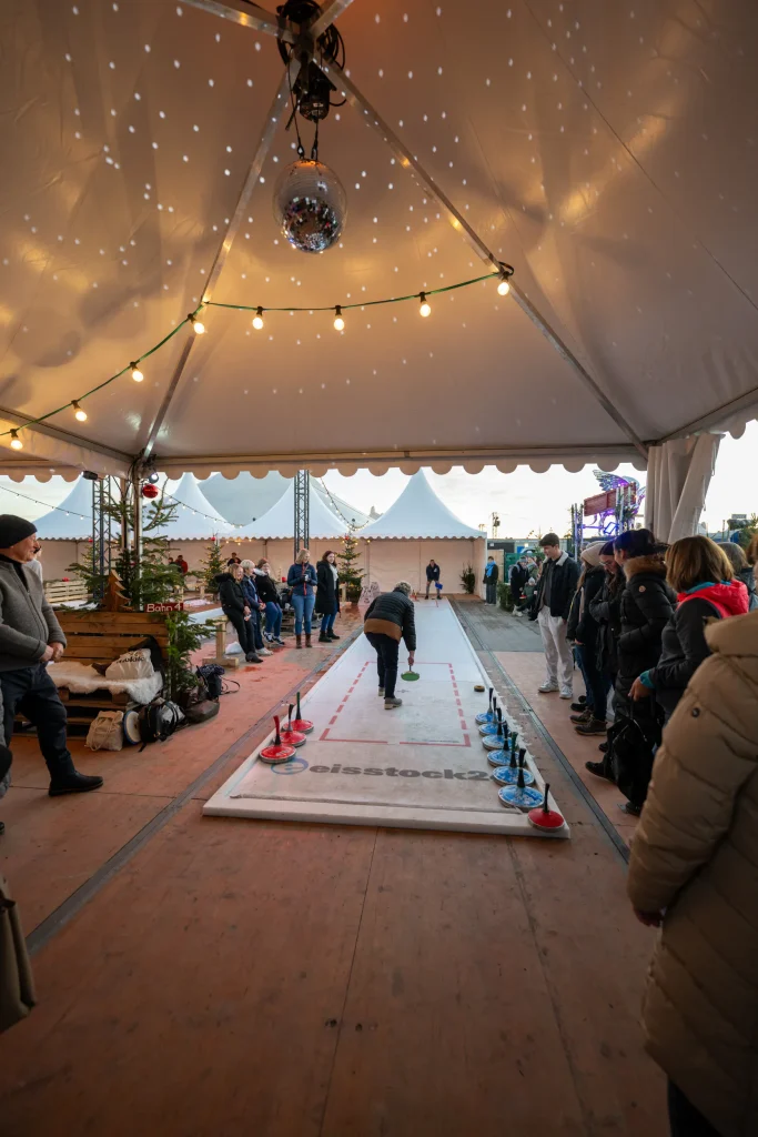 TwWi24_241127_c_alexander_scharf-134 - IN München - Das Stadtmagazin People watch a curling-like game being played on a long white lane inside a festival tent, with string lights and a disco ball overhead.
