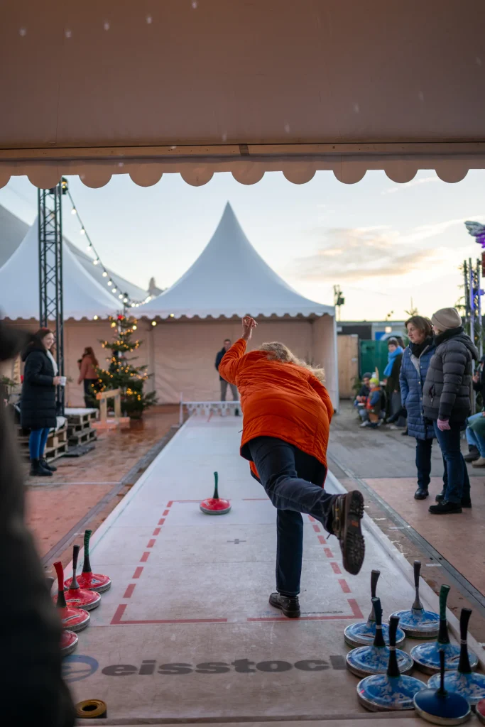 TwWi24_241127_c_alexander_scharf-144 - IN München - Das Stadtmagazin Person in an orange jacket throws a curling stone along an outdoor rink as spectators watch under white tents nearby.