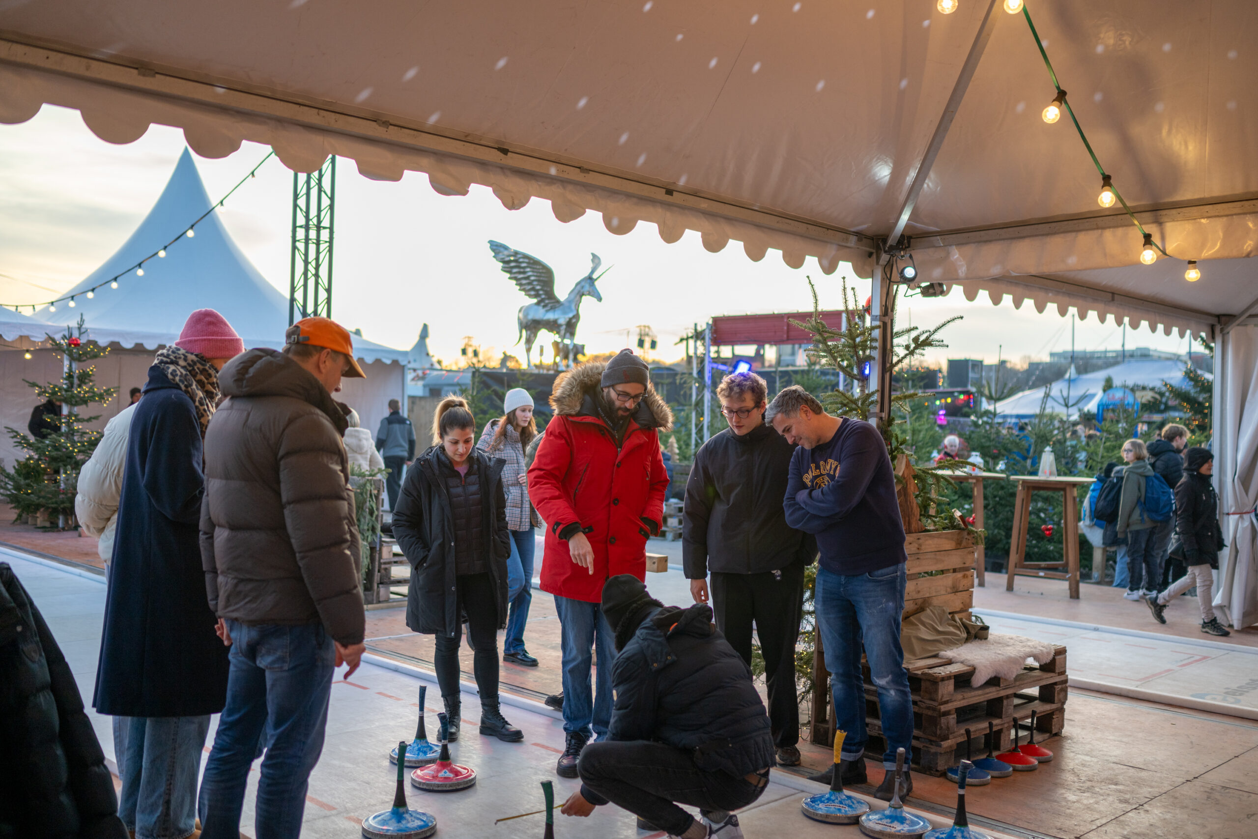 TwWi24_241217_c_alexander_scharf-152 - IN München - Das Stadtmagazin People in warm coats gathered under a white event tent watching a curling game on an ice rink, with curling stones lined up nearby.