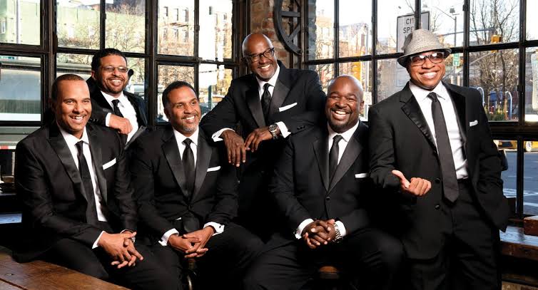 Six men in black suits pose together for a group photo in a glass-walled indoor space, smiling at the camera.