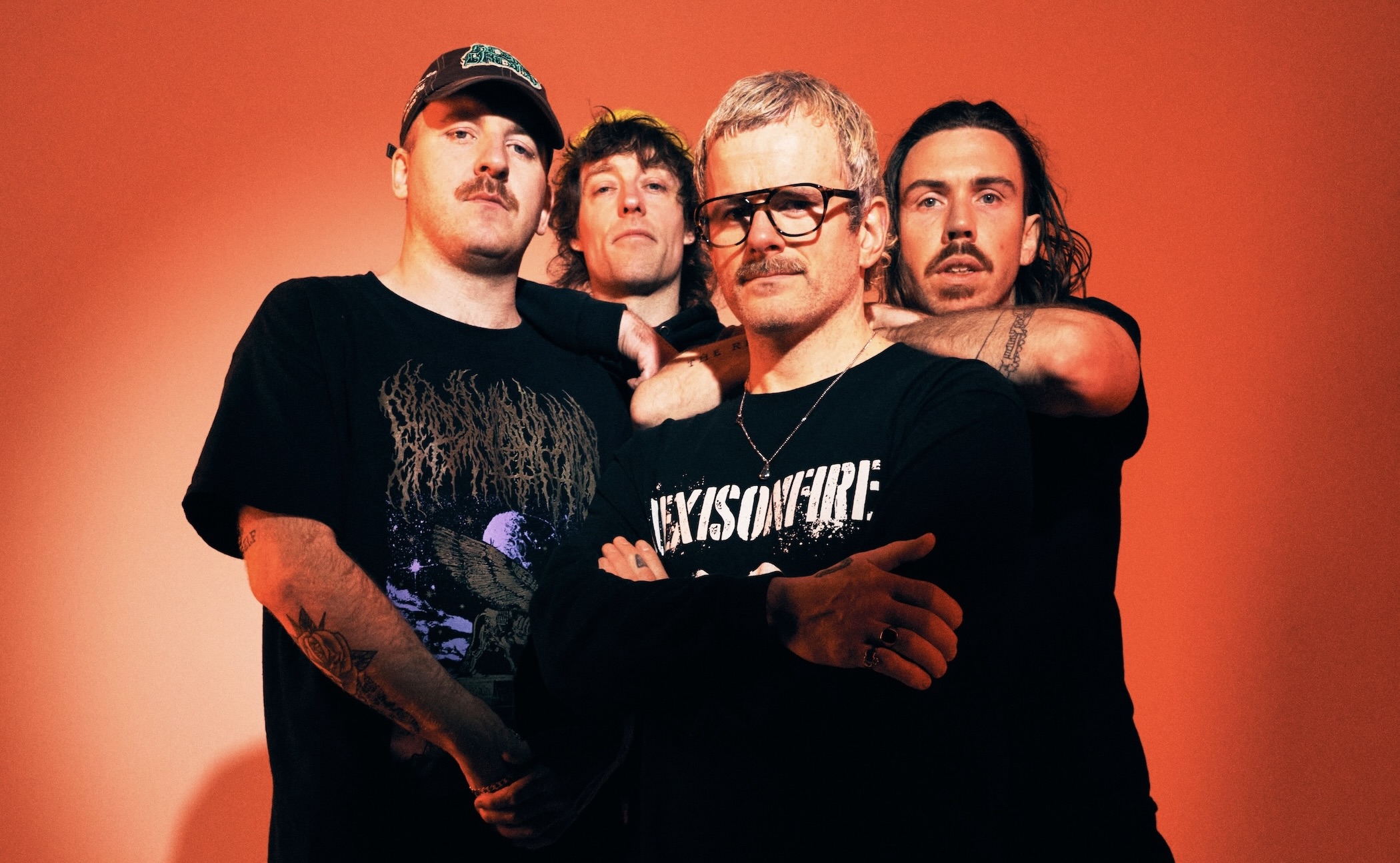 Four men in black shirts pose closely together against an orange background, arms around each other.