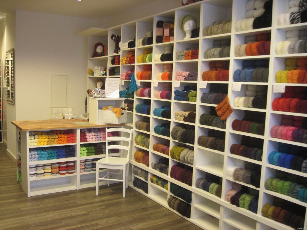 Wollstudio_Laim - IN München - Das Stadtmagazin Yarn shop interior with white cubby shelves filled with colorful skeins arranged by color, a wooden counter, and a white chair in the foreground.