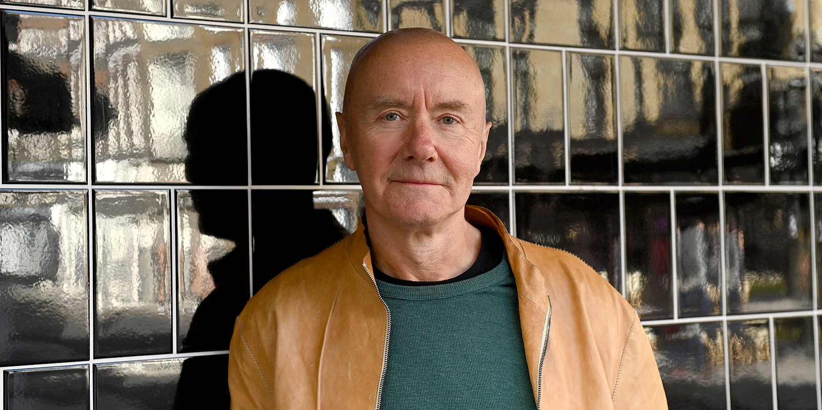 Portrait of a bald, middle‑aged man in a green sweater and tan jacket, standing against a glossy tiled wall with a neutral expression.
