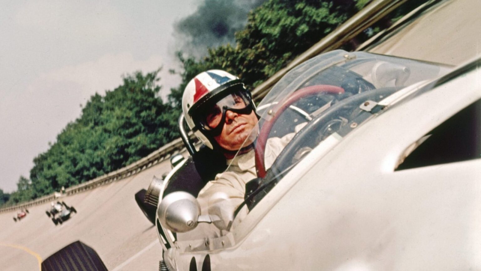 Driver in a vintage open-cockpit race car, wearing a striped helmet and goggles, on a curving track with trees in the background.