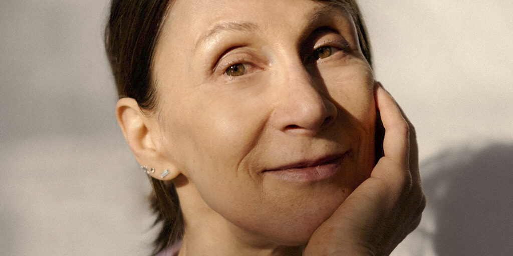 Martina Hefter - IN München - Das Stadtmagazin Close-up of a smiling woman with short dark hair touching her cheek with a hand, wearing small earrings.