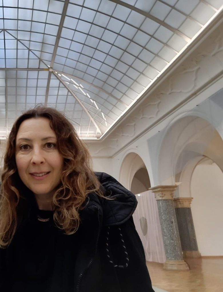 Woman with wavy brown hair in a black coat stands in a bright interior with a glass skylight and ornamental arches.