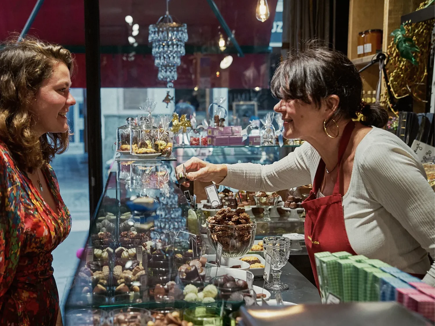 mucho-munich-chocolate-festival-schokoladenverkostung-confiserie-stand - IN München - Das Stadtmagazin Shopkeeper in a red apron hands a wrapped treat to a customer at a glass display filled with chocolates and pastries.