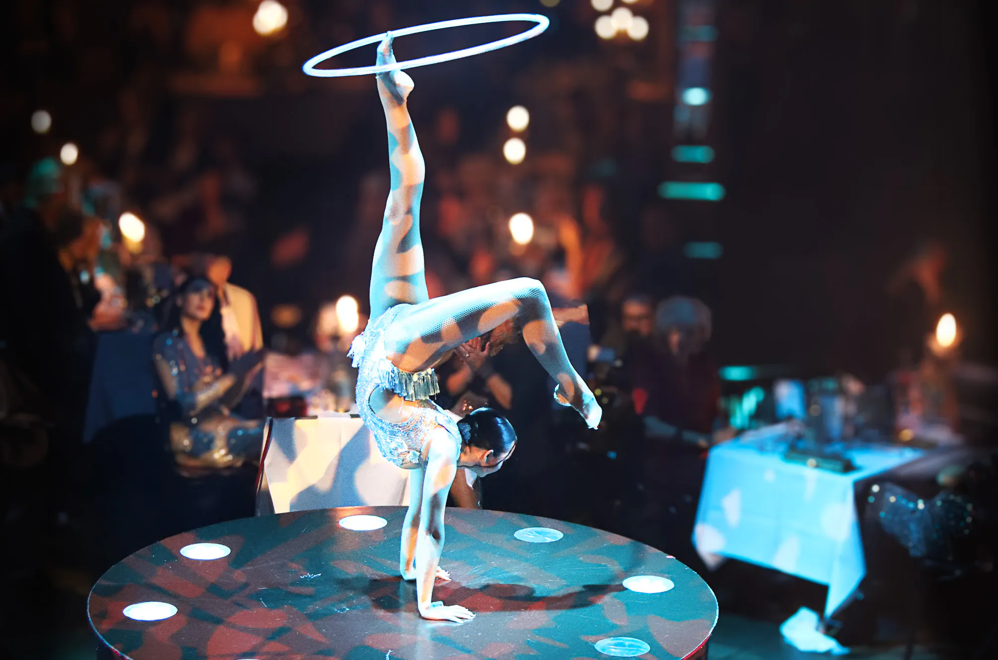 Acrobat balances on a circular stage, leg raised overhead with a glowing hoop, audience blurred in the background.