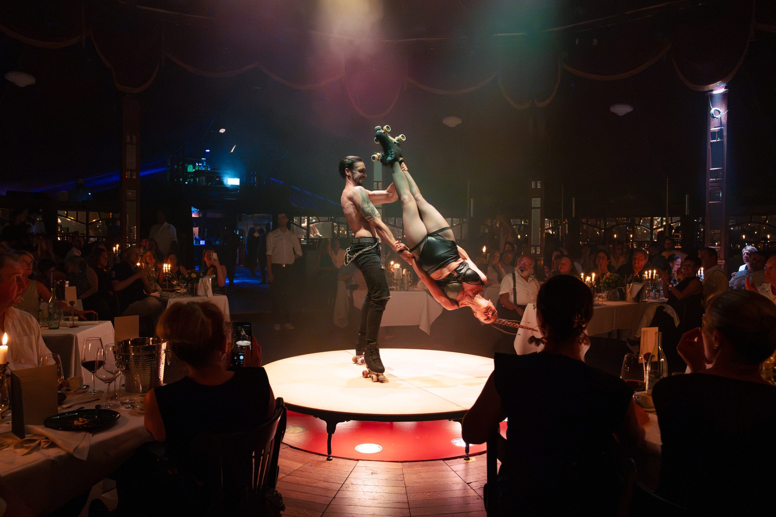 A shirtless male trapeze/roller-skating performer lifts a female performer in fishnet tights on a circular stage amid a dimly lit restaurant audience. Alternative: two skaters balance a high pose on a lit circular platform with diners around.