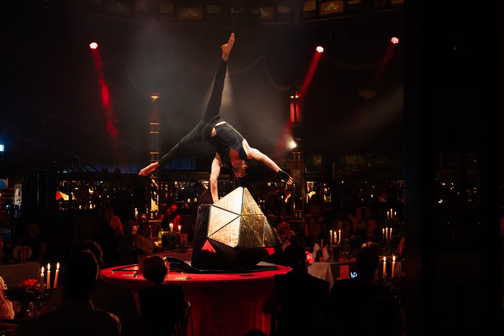teatro_SommerSeason26-22 - IN München - Das Stadtmagazin Acrobat balances on a geometric prop atop a red-lit stage, arms spread, while diners watch in a dim, candlelit venue.