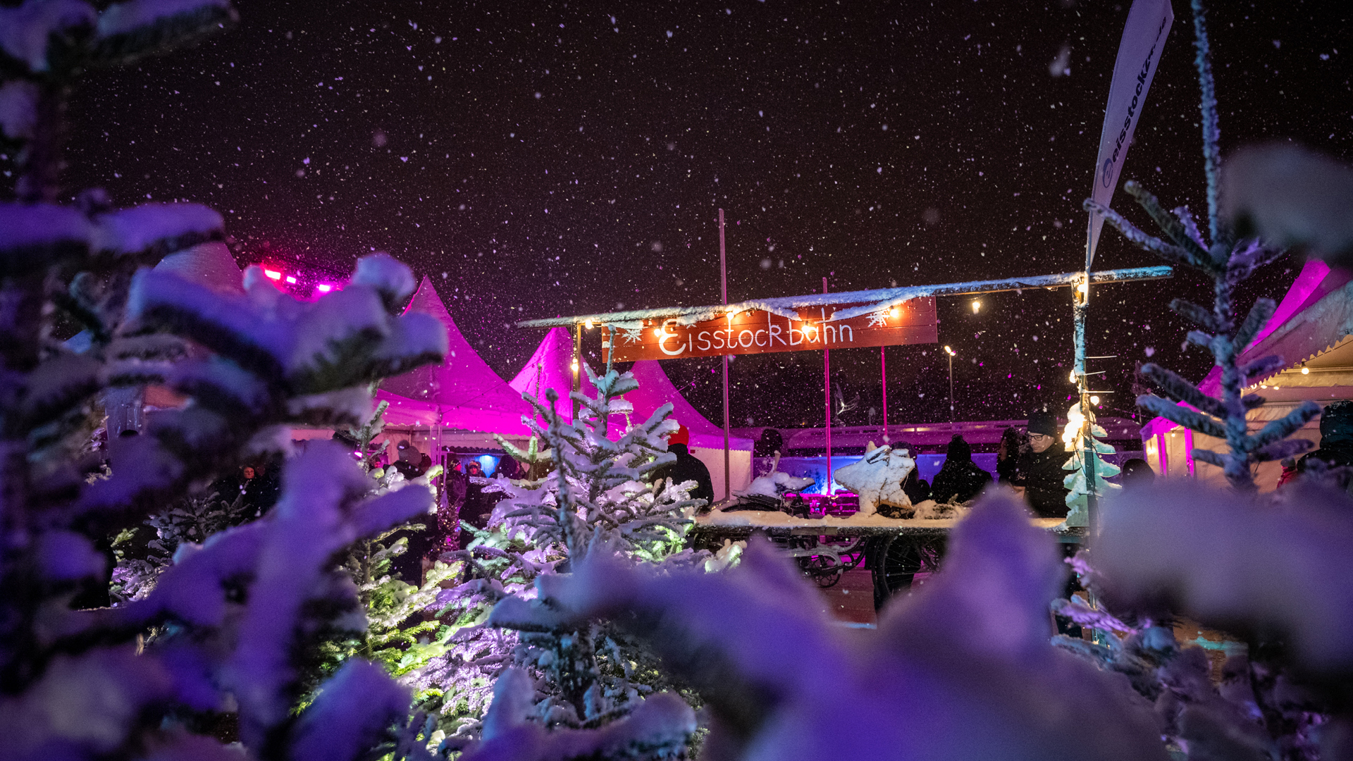 Snowy outdoor festival at night with purple and warm lights, banner reading Eisstockbahn, tents, and people.