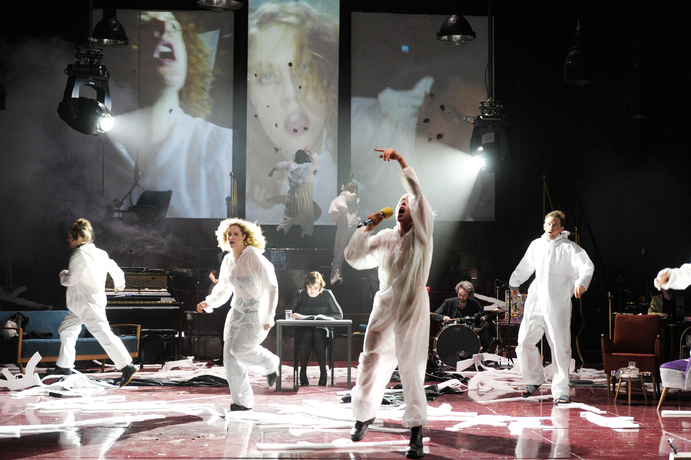 Jelinek - IN München - Das Stadtmagazin Stage performance with dancers in white outfits, musicians, and large video projections of faces in the background.